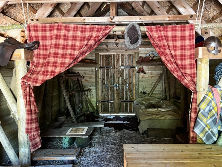 Picture shows inside the longhouse at martin mere. There is a bed to the right, a cow skin rug on the floor, and various pieces of furniture around.